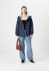 Giacca impermeabile blu navy con interno grigio, polsini elasticizzati e orlo con cordino, abbinata a jeans a gamba larga blu chiaro e sneakers nere.