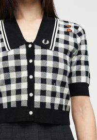 Cardigan à manches courtes à carreaux noirs et blancs avec boutons blancs, col noir avec rayures blanches, patchs en forme de cœur rouges sur l'épaule, porté par une personne.