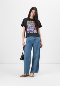 Maglietta grafica nera con un design colorato "Oasis", abbinata a jeans blu larghi. Gli accessori includono una piccola borsa nera e scarpe beige.
