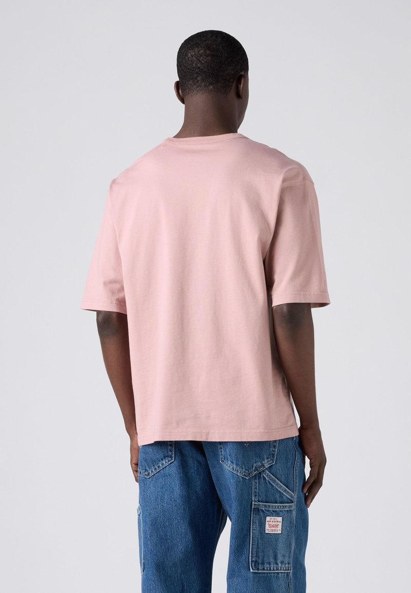 Homme debout, vu de dos, portant un t-shirt manches courtes rose clair uni et un pantalon cargo en denim bleu avec poches visibles et étiquette.