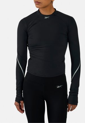 Mujer con camiseta deportiva negra ajustada de manga larga y mallas con logotipos reflectantes de Reebok en el pecho, las mangas y la cadera.