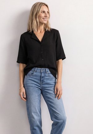 Kvinde med blondt hår iført en sort skjorte med knapper og lyseblå jeans står smilende og læner sig op ad en hvid væg.