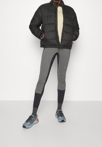 Chaqueta negra con volumen, camisa de un tono beige claro, leggings grises con acentos en azul marino y zapatos deportivos azules con cordones negros y detalles en naranja.