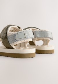 Sandales avec une semelle beige, des lanières grises et des accents olive. Elles disposent de fermetures réglables et d'un tissu texturé. Semelle en caoutchouc antidérapante.