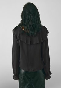 Donna con capelli ricci e scuri indossa una blusa nera a maniche lunghe con colletto e polsini arricciati, abbinata a una gonna in pelle verde scuro.