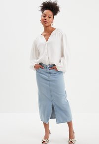 Camisa blanca texturizada de botones con mangas relajadas, combinada con una falda de mezclilla azul clara hasta la rodilla con una abertura frontal. Sandalias plateadas.