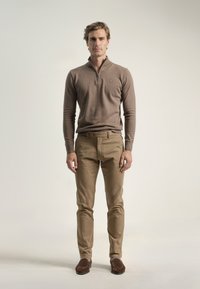 Jersey marrón de cremallera, chinos beige y zapatos marrones oscuros. El conjunto presenta un diseño ajustado, una textura suave y detalles sutiles.
