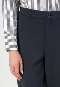 Marinebukser laget av et glat og elastisk stoff. Har en synlig midjebånd, knappeåpning og frontlomme. Bært med en svart og hvit stripet skjorte.