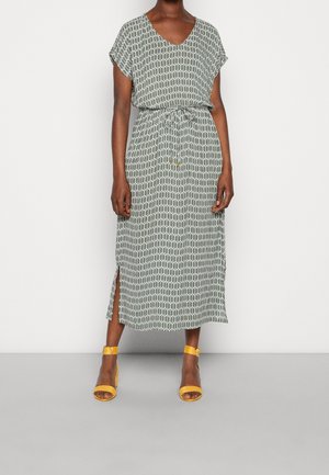 Osoba o ciemnej karnacji ubrana w wzorzystą sukienkę midi z krótkimi rękawami i paskiem w talii, w połączeniu z żółtymi sandałami na blokowym obcasie z paskiem wokół kostki.