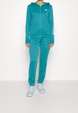Personne portant un survêtement Adidas bleu sarcelle avec un sweat à capuche zippé et un pantalon de jogging assorti, associé à des baskets bleu clair et blanches.