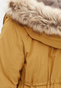 Žlutý polstrovaný kabát s hnědým kožichovým lemem na kapuci, s elastickým páskem a strukturovanou tkaninou. Vhodné do chladného počasí.
