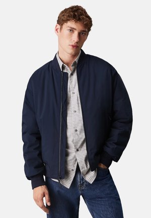 Chaqueta bomber navy con puños y dobladillo acanalados, tela suave, cremallera frontal, superpuesta a una camisa gris de cuadros, combinada con jeans azules.