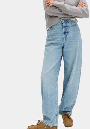 Person iført lyseblå viddeben jeans, grå sweater og brune ruskindsmokkasiner, stående med armene krydset.