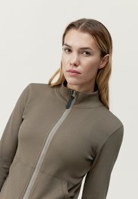 Giacca aderente a coste con zip in verde oliva, dotata di un collo alto e zip a contrasto con un tirante. Tessuto liscio, maniche lunghe.