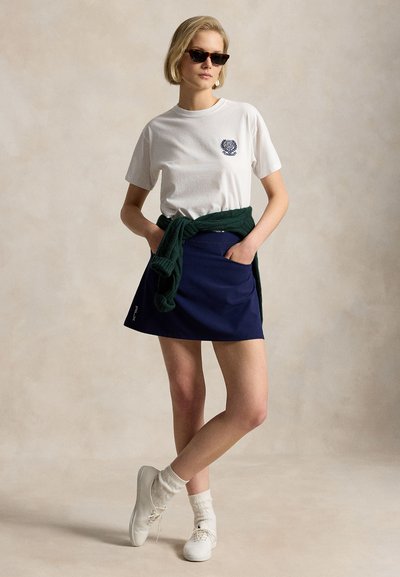 Γυναίκα που φοράει λευκό t-shirt, μπλε ναυτικό φούστα, λευκά αθλητικά και γυαλιά ηλίου, με πράσινο πουλόβερ δεμένο γύρω από τη μέση της, στέκεται μπροστά σε ουδέτερο φόντο.