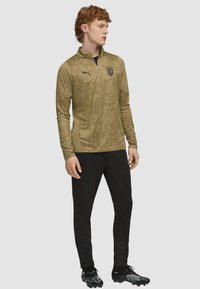 Olivengrøn atletisk zip-up trøje med struktureret mønster, parret med sorte bukser og sorte sportssko. Har et logo på brystet.
