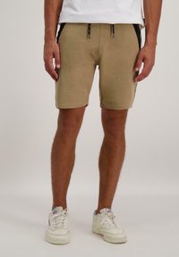 Lichtbruine shorts gemaakt van zacht materiaal, met zwarte zijaccenten, verstelbare trekkoorden en een casual pasvorm. Draagt met witte sneakers.