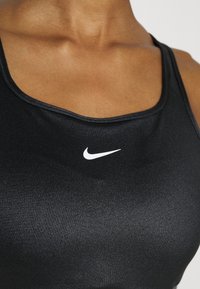 Soutien-gorge de sport noir en tissu lisse et élastique, avec un col arrondi et un petit logo Nike blanc sur la zone gauche de la poitrine.