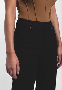 Jeans de mezclilla negra de cintura alta con una textura suave, con herrajes de cobre y bolsillos frontales, combinados con una blusa marrón de canalé.