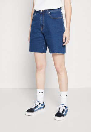 Persoon draagt donkerblauwe spijkershorts van middelhoge lengte, witte Nike sokken en blauwe en zwarte sneakers, staand tegen een effen witte achtergrond.