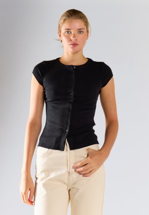 Giovane donna con capelli biondi che indossa una maglietta nera aderente con bottoni e maniche corte e pantaloni beige chiaro a vita alta, in piedi davanti a uno sfondo grigio.