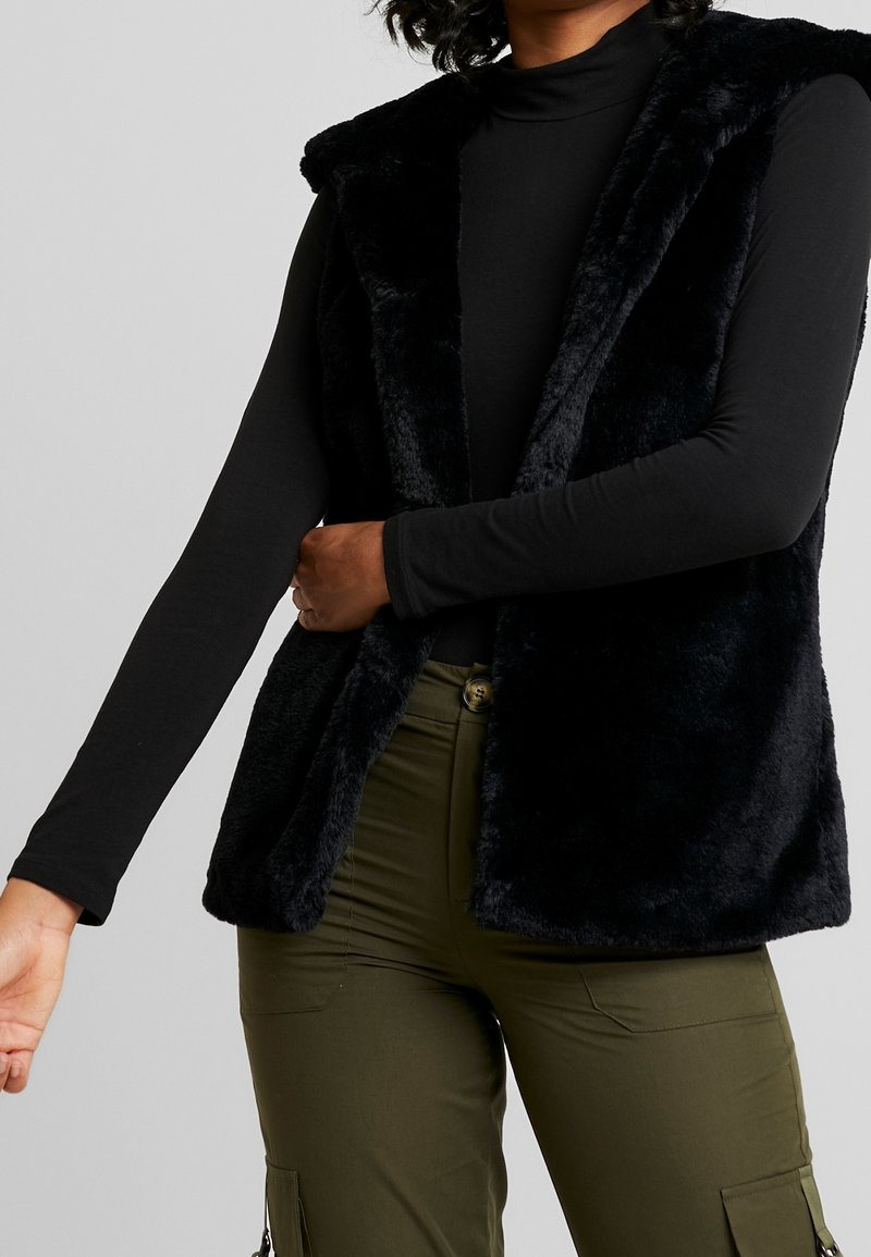 Personne portant un gilet en fausse fourrure noire sur un haut à manches longues noir et un pantalon cargo vert olive, debout avec un bras tendu et une main touchant le gilet.