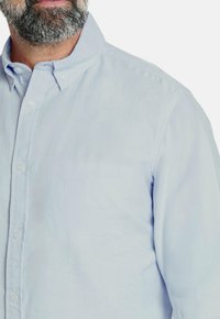 Hombre con barba que lleva una camisa de botonadura ligera azul con cuello abotonado contra un fondo blanco liso.