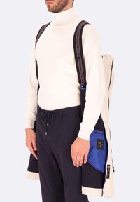 Pullover bianco a collo alto, pantaloni scuri e giacca multifmateriale con foderatura blu e accenti in contrasto; presenta cerniere e cinturini con logo.