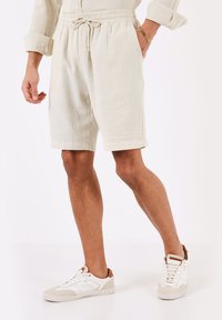 Pantaloni corti beige chiaro in tessuto testurizzato, con cintura elastica e tasche laterali, abbinati a sneakers bianche e beige.
