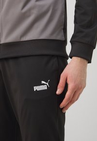 Črne športne hlače z belim logotipom Puma na spodnjem levem delu. Tkanina je gladka z rahlim sijajem, manšete pa so rebraste.