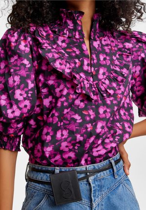 Kvinde iført en bluse med puffede ærmer og pink blomsterprint samt flæsekrave, stukket ned i højtaljede blå jeans med en sort bæltepose.