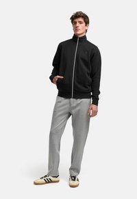 Svart hettejakke med høy krage, grå joggebukser og beige joggesko med svarte striper. Modellen står med hendene i lommer.