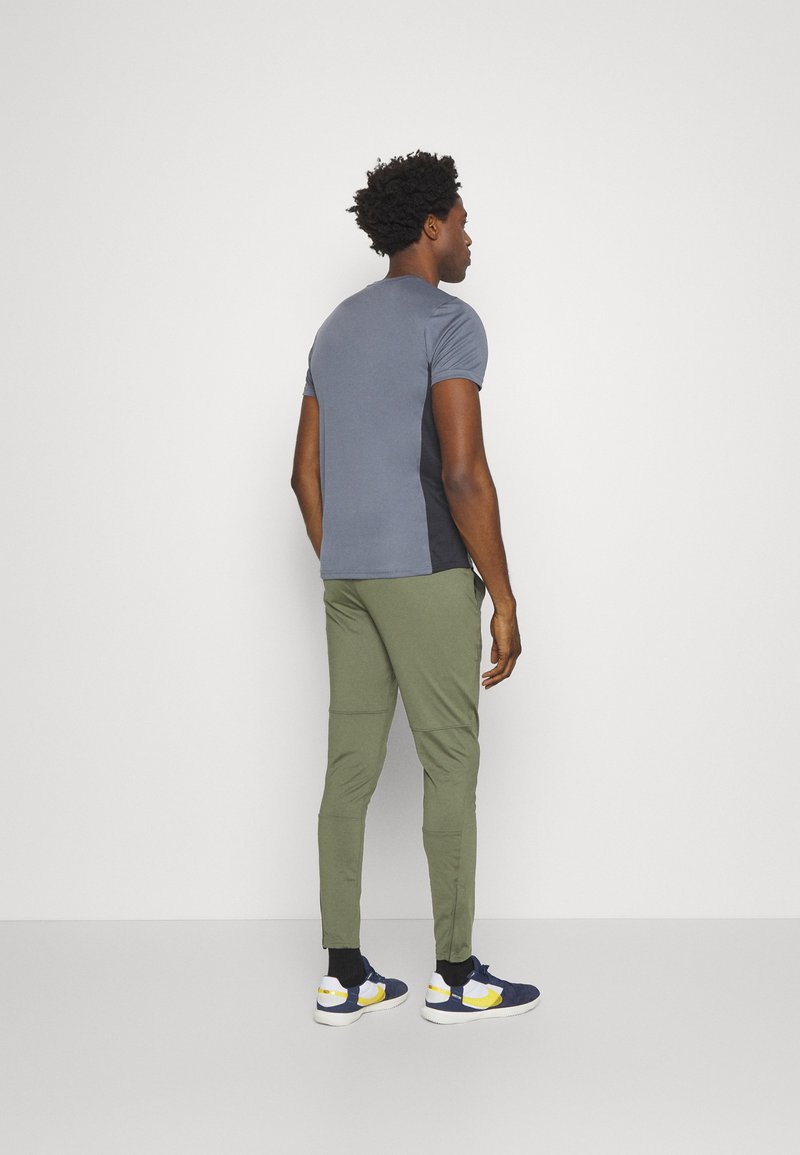 Homme debout face à un mur, portant une chemise gris à manches courtes, un pantalon vert olive, des chaussettes noires et des baskets bleu marine avec des accents jaunes.