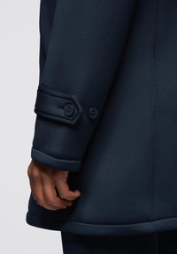 Abrigo azul marino con diseño entallado, detalle de puños abotonados y textura suave. Presencia de costuras visibles y un dobladillo moldeado.