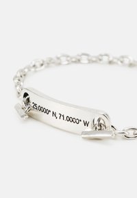 Bracelete de metal prateado com um design de corrente, apresentando uma placa gravada que exibe as coordenadas: "25.0000° N, 71.0000° W."