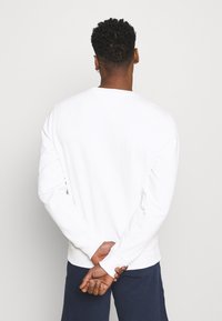 Felpa bianca a maniche lunghe con girocollo, dotata di polsini e orlo a coste, mostrata da dietro, indossata con pantaloni blu navy.