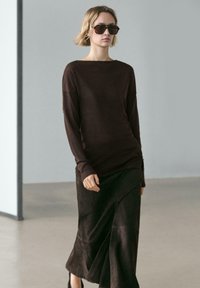 Brązowa bluzka z długim rękawem z miękkiej tkaniny, zestawiona z ciemnobrązową zamszową spódnicą z bocznym rozcięciem. Całość uzupełniają czarne, oversize okulary przeciwsłoneczne.