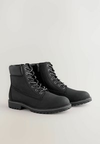 Ein Paar schwarze Schnürstiefel mit gepolstertem Kragen und robusten Sohlen auf einem schlichten hellen Hintergrund.