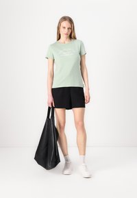 T-shirt en coton vert menthe à manches courtes avec texte blanc, associé à un short noir et un grand sac fourre-tout en cuir noir.