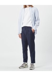 Camicia azzurra a righe con bottoni sopra una maglietta bianca, abbinata a pantaloni plissettati blu navy e scarpe sportive grigie.