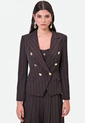 Femme portant un blazer croisé à fines rayures foncé avec des boutons dorés et un pantalon assorti, debout devant un fond clair uni.