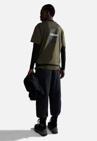 T-shirt in cotone verde oliva con una grafica circolare sul retro, abbinato a pantaloni blu navy scuri e stivali neri. Persona che tiene un maglione scuro.