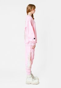 Sudadera rosa y pantalones de chándal con diseño de alas bordado. La sudadera tiene un corte relajado; los pantalones cuentan con puños acanalados. Llevado con botas blancas.