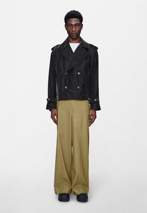 Mladi moški stoji, oblečen v črno dvoredno jakno, bež široke hlače in črne čevlje, na enobarvnem belem ozadju.