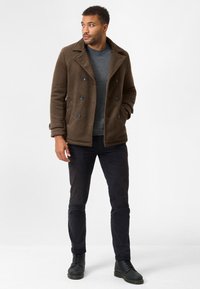 Brown textured double-breasted coat with large buttons over a gray sweater, black slim-fit pants, and black lace-up boots.