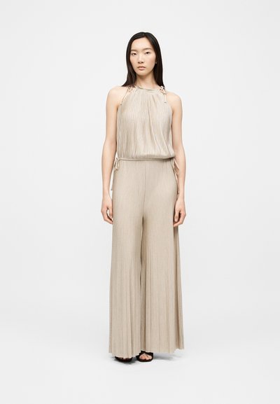 Femme asiatique portant un top sans manches plissé beige à col halter et un pantalon large assorti, debout devant un fond blanc uni.