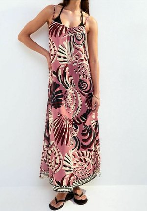 Femme portant une robe longue rose avec des motifs abstraits de coquillages en noir, blanc et marron, bretelles fines, et sandales noires, debout contre un mur blanc.
