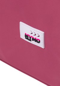 Borsa in tessuto rosa con un'etichetta bianca prominente con scritte nere e rosa e dettagli in rosa, che presenta una trama liscia e un design semplice.