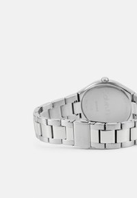 Reloj de acero inoxidable plateado con una pulsera de eslabones, caja redonda y nombre de la marca grabado en la parte posterior. Textura suave y pulida.
