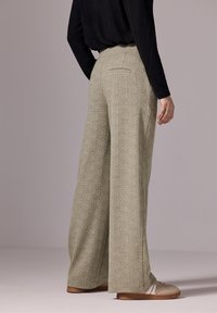 Pantalones de pierna ancha en una tela texturada de color beige claro. Tienen un bolsillo trasero y se combinan con una blusa negra y zapatos de tonos neutros.